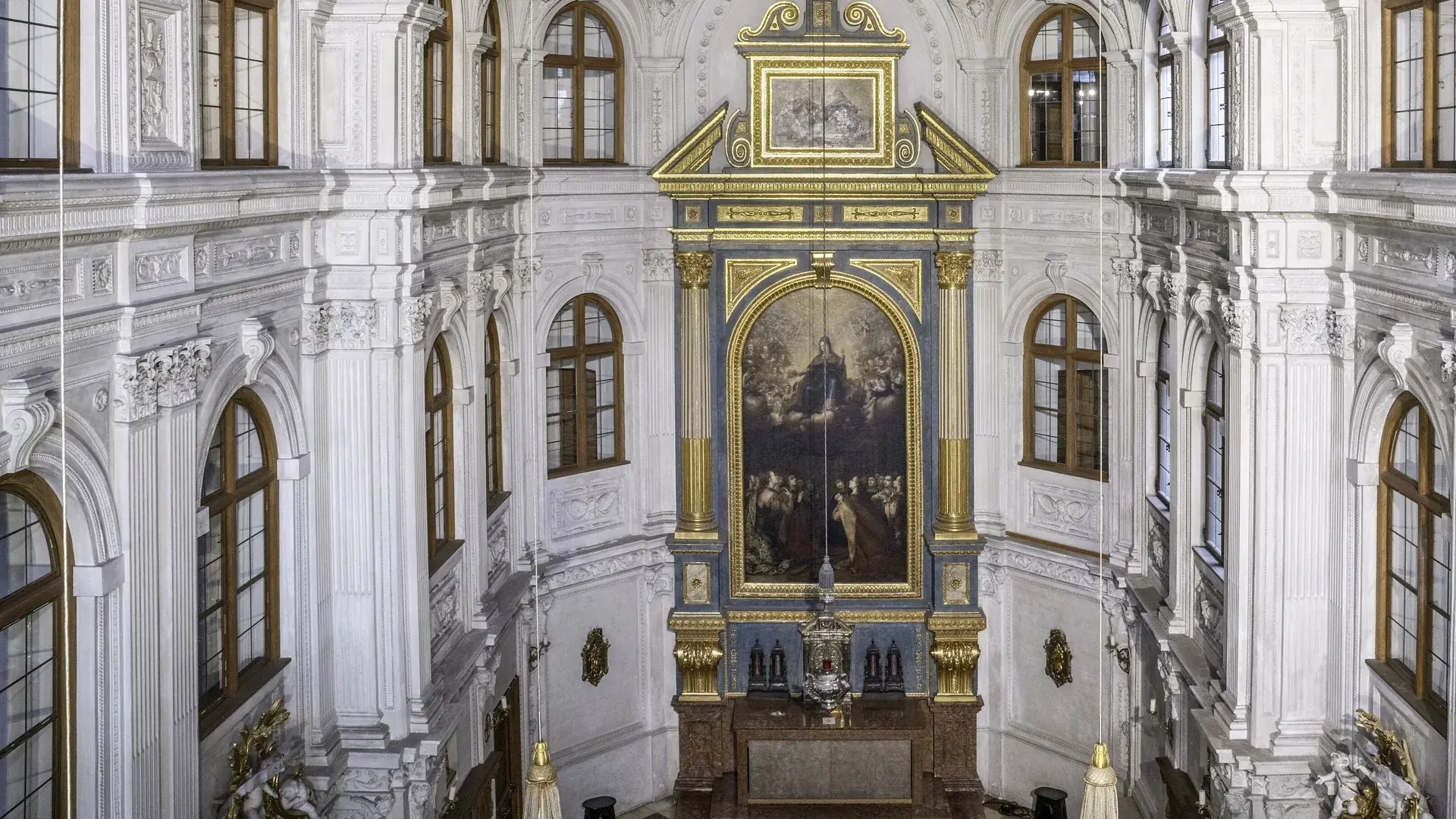 Pohled dolů do dvorní kaple mnichovské Rezidence: bílé štukové stěny, vysoká klenutá okna s hnědými rámy a pozlacený rám oltáře kolem velkého obrazu Panny Marie ve slávě.