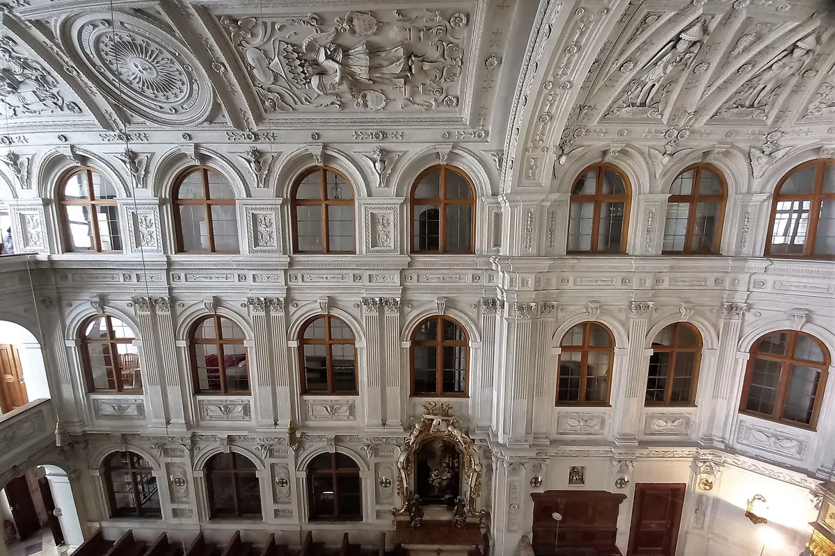 Horní stěna kaple se dvěma patry obloukových oken, štukovými panely a dřevěnými lavicemi pod nimi.