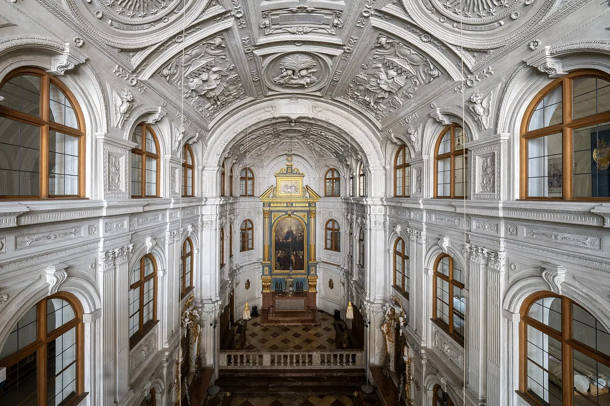 Dvorní kaple pohledem z galerie, s vysokou bílou štukovou lodí, obloukovými okny s hnědými rámy a pozlaceným oltářem v zadní části.